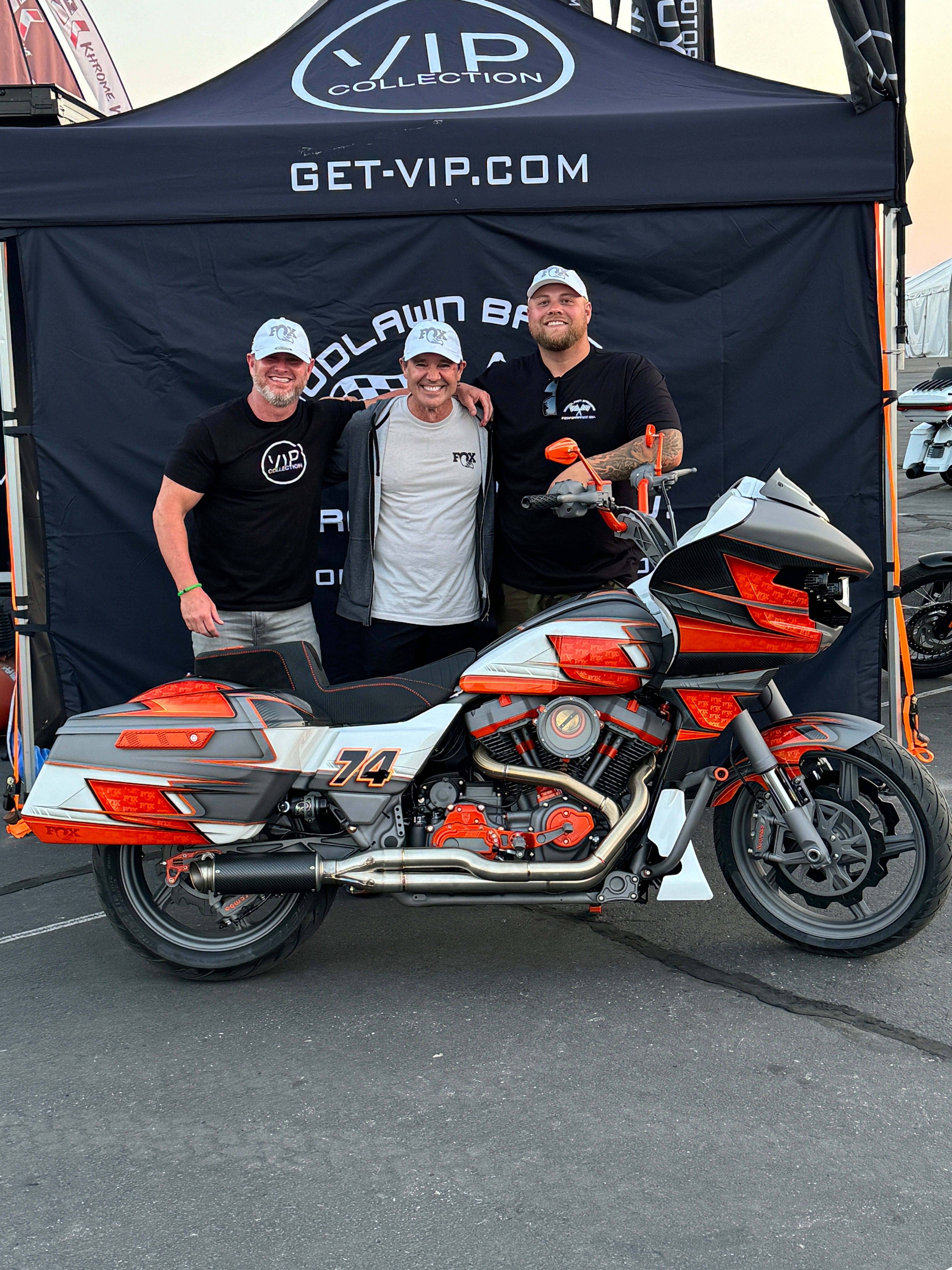 Three men standing in front of a black VIP Collection canopy tent, with a large black, orange, and white motorcycle parked in the foreground on asphalt.