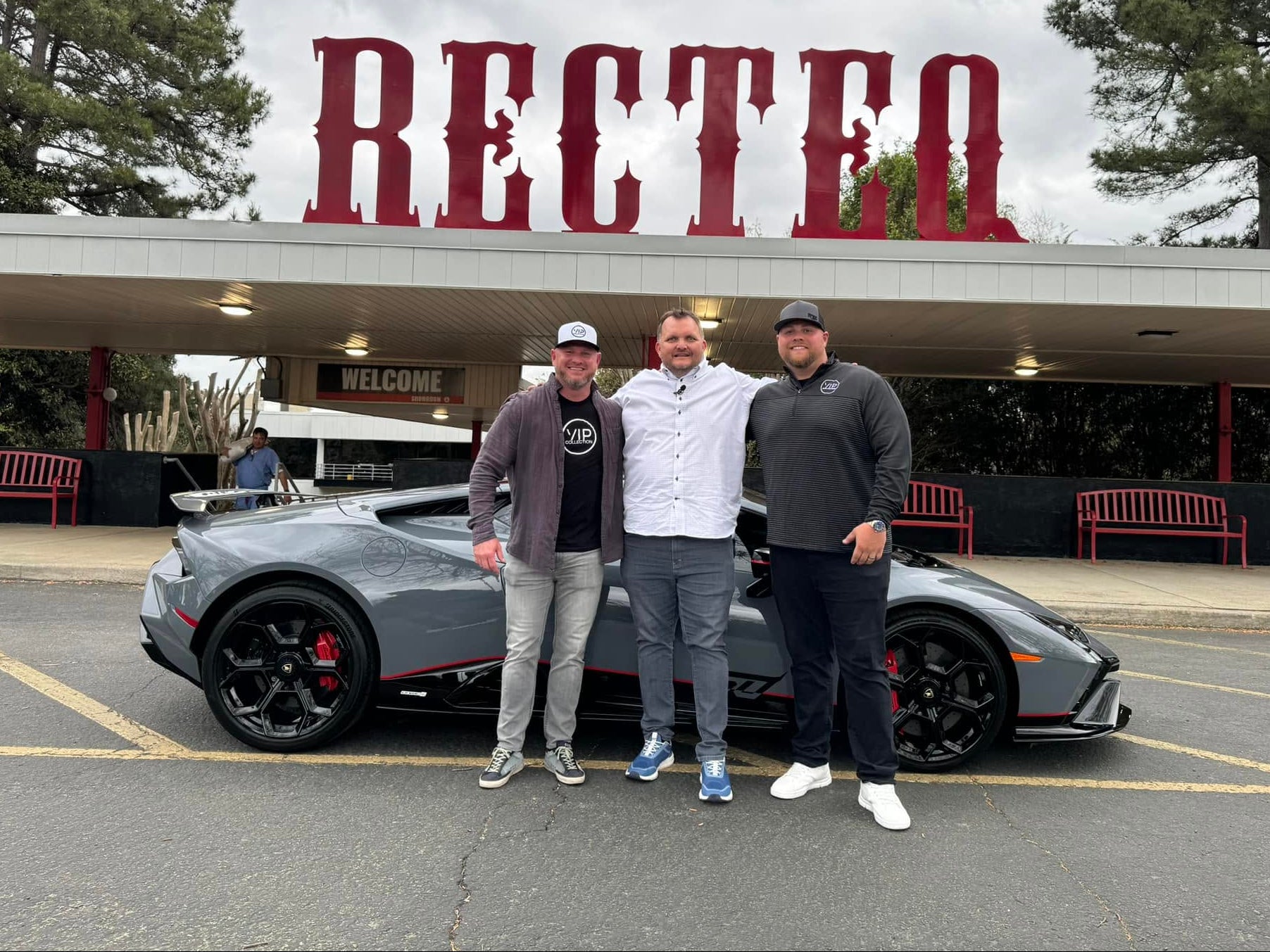 Three men standing in front of a gray sports car parked outside a building with a large red 'RECTEQ' sign overhead.